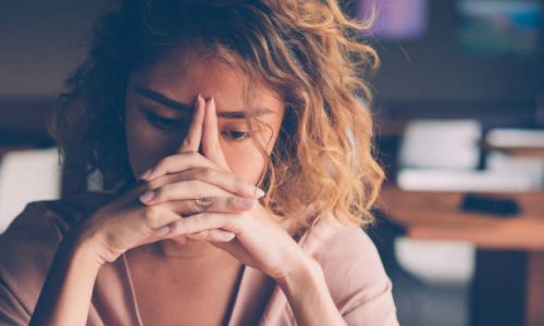 Closeup of sad young Asian woman at cafe leaning head on clasped hands and staring into vacancy. Tired freelancer feeling burnout. Stress and bad news concept